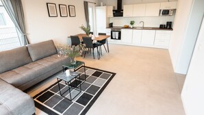 Una sala de estar moderna con un sofá gris, una mesa de centro y un comedor con sillas y una mesa. Se ve una cocina con gabinetes y electrodomésticos blancos al fondo.