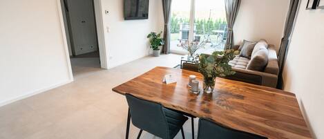 Un espace repas moderne avec une table en bois, deux chaises, une plante en pot et une vue sur un jardin grâce à de grandes fenêtres.