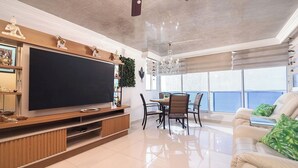 Ein geräumiges Wohnzimmer mit großem Flachbildfernseher, eine Essecke mit Tisch und Stühlen sowie ein Balkon mit Blick.