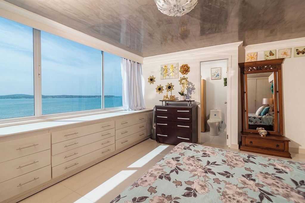 A bedroom with a large window, a bed with a floral bedspread, a dresser, a mirror, and wall art.