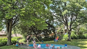 A playground with a slide, swings, and a small pool area surrounded by grass and trees.