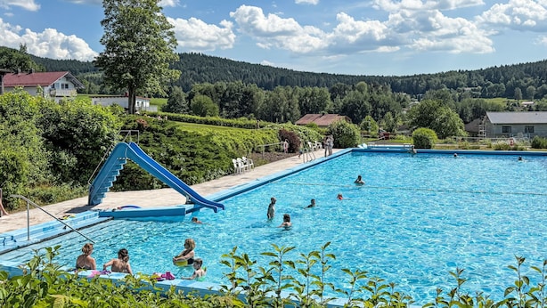 A swimming pool with a slide, surrounded by greenery and houses, under a clear sky.