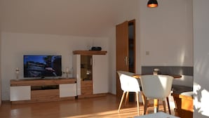 A modern living room with a TV, wooden entertainment unit, white chairs, and a dining table.