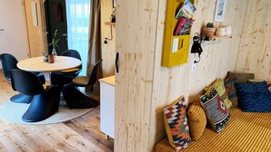 Una camera accogliente con pavimento in legno, un tavolo rotondo con sedie, un divano con cuscini decorati, uno scaffale giallo con libri e una pianta in vaso, e una finestra con tende.