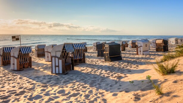 Une plage bordée de rangées de chaises de plage et de petits abris, dont certains sont surmontés de toiles rayées, sous un ciel dégagé.