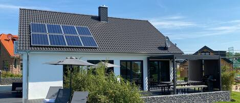 A modern house with a sloped roof, solar panels, a patio with lounge chairs, and a stone wall.