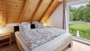 Une chambre avec un plafond en pente, une grande fenêtre et un lit recouvert d’une literie à motifs.