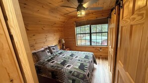 Une chambre dans un chalet en bois, avec un lit, un ventilateur de plafond, une fenêtre et une table de chevet.
