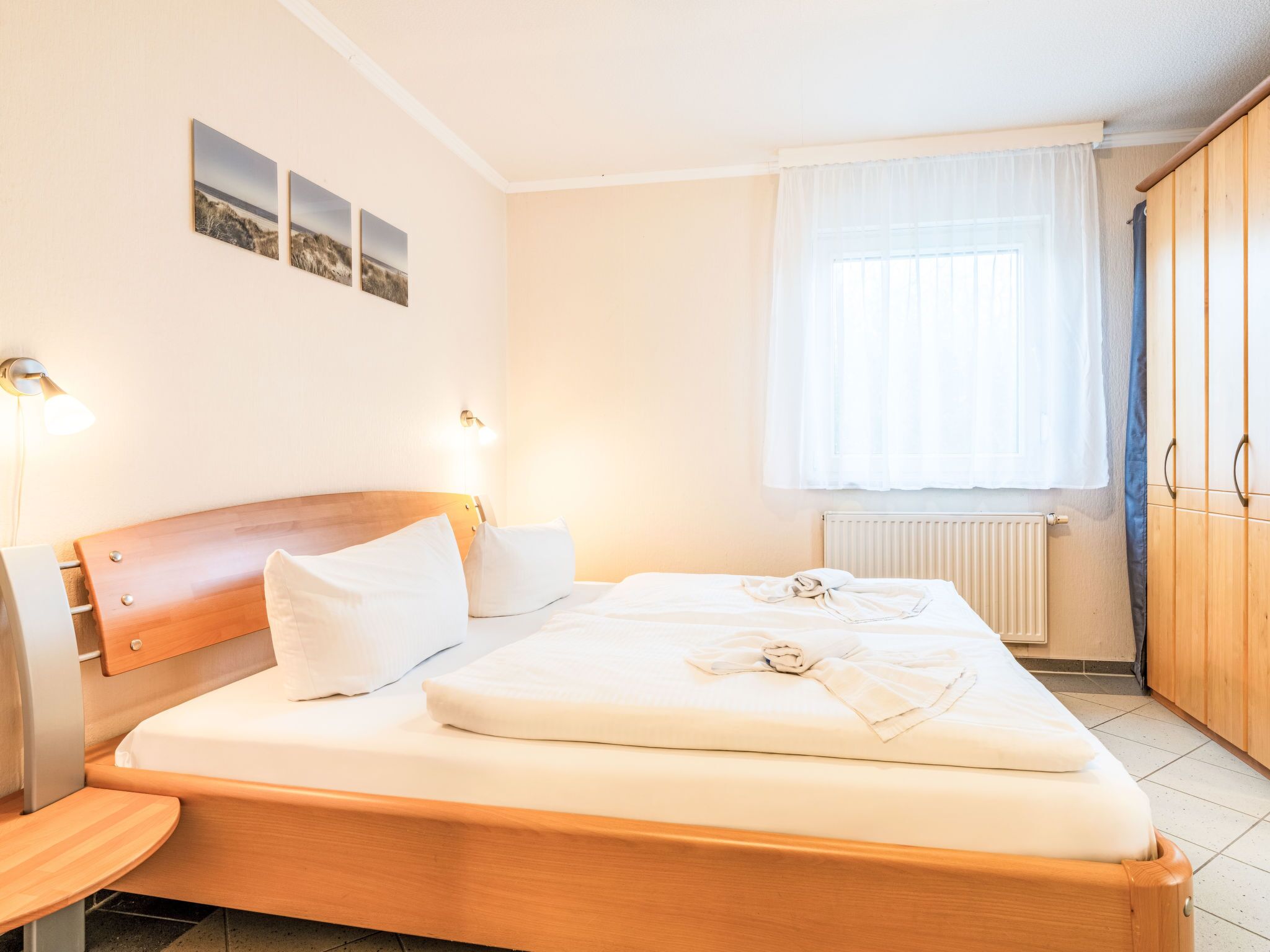 A bedroom with a wooden bed, white bedding, a nightstand, a wall-mounted lamp, a window with curtains, and a radiator.