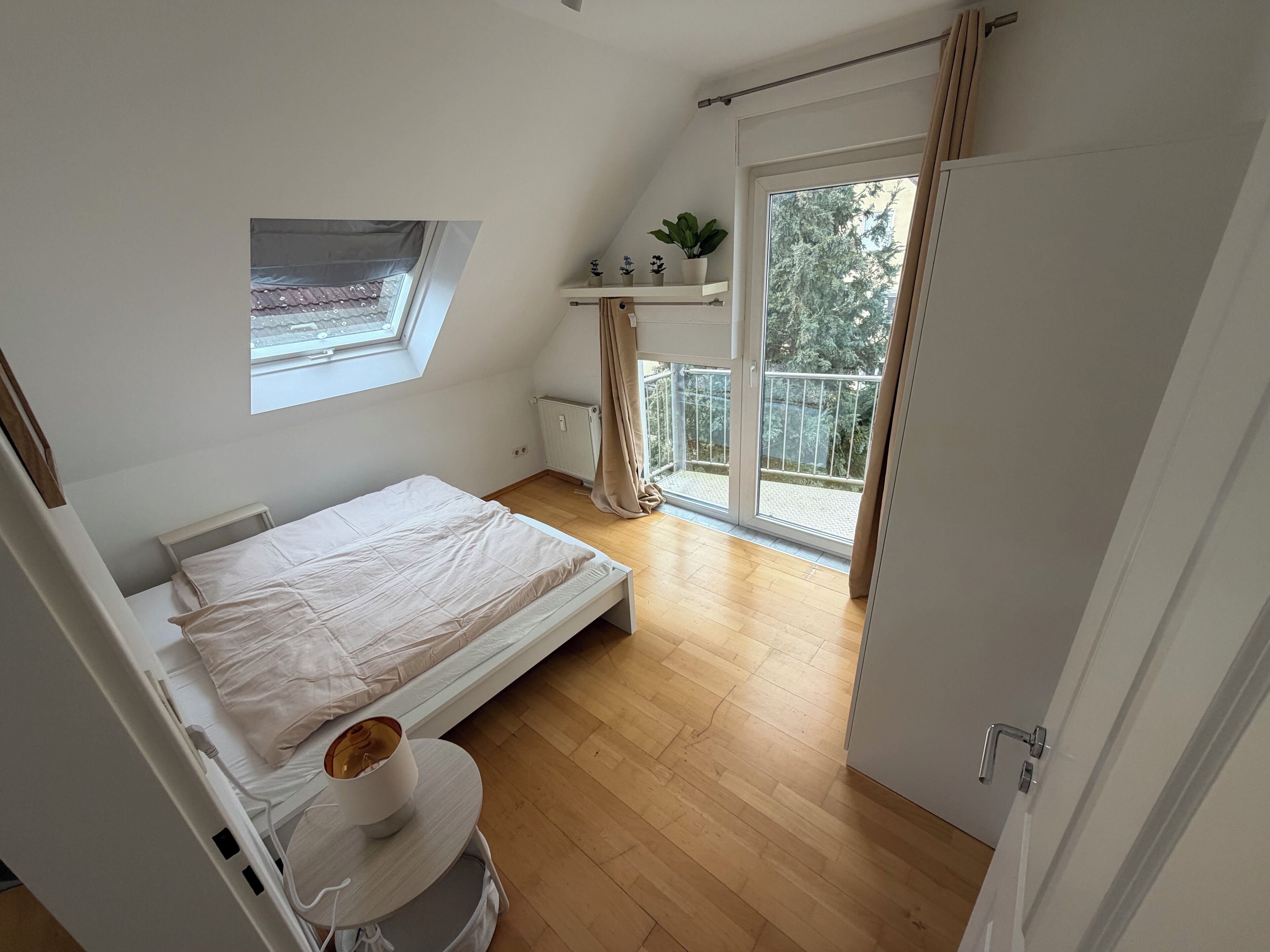 Une chambre avec un lit simple, une petite fenêtre, un radiateur et un balcon donnant sur des arbres.