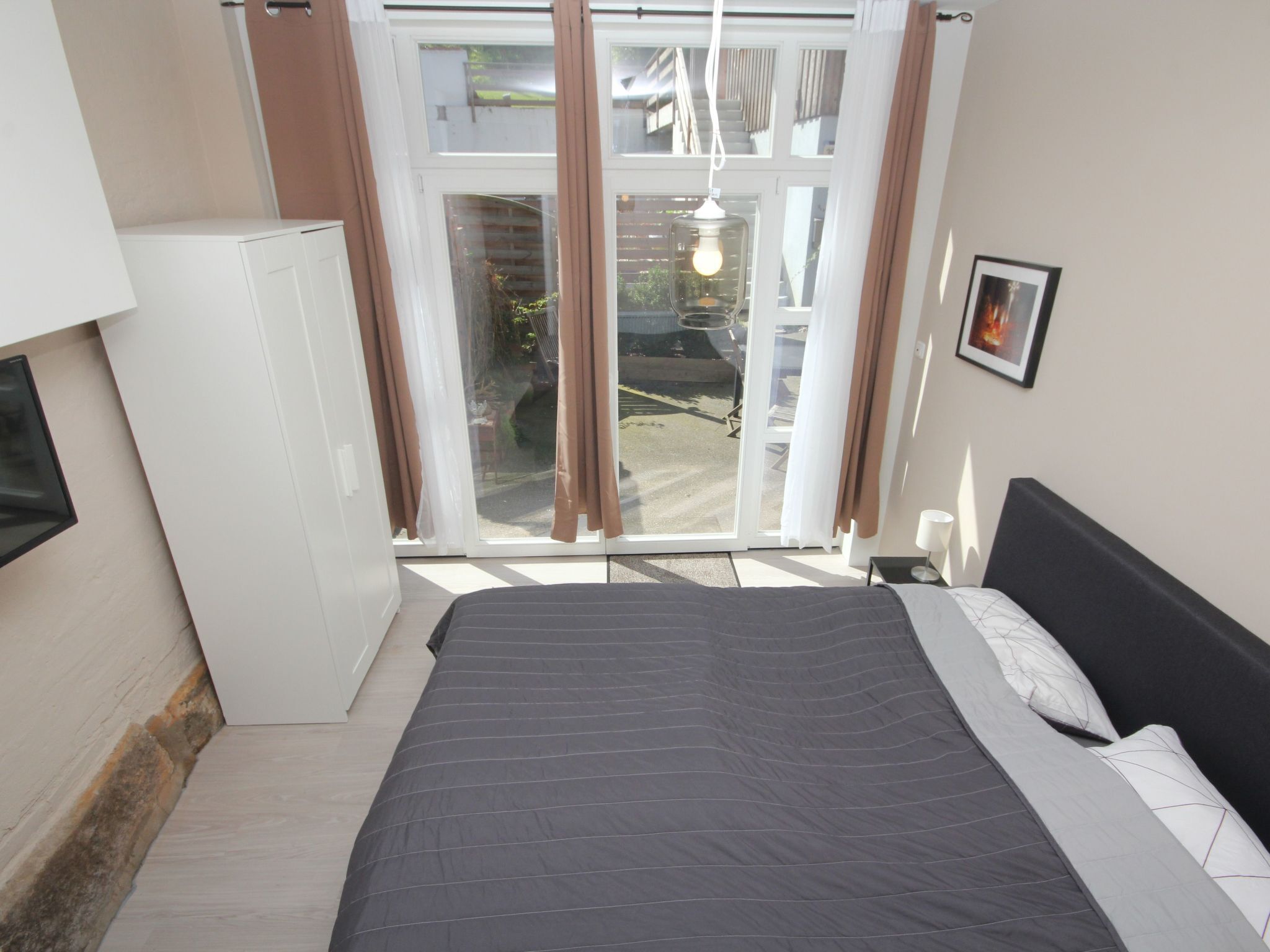 A bedroom with a bed, a small white cabinet, a sliding glass door to the outside, and a framed picture on the wall.