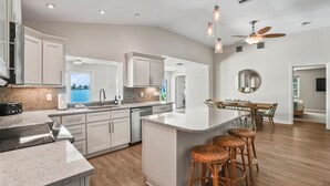 A modern kitchen with white cabinets, a large island, and a dining area with a round table and wicker bar stools. - Waterfront 4 BR Home, Pool, Private Dock, Kayaks, Putting Green, Walk to Beach (Indian Rocks Beach)