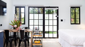 A modern room with a bed, a dining table with chairs, a small cart with bottles, and large windows with black frames. - Altavida Lodge · New Studio with Pool in El Valle · N5 (Valle de Anton)