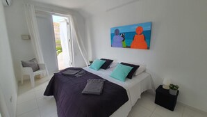 Ein Schlafzimmer mit einem Bett, Kissen, einem Deckenventilator, einem Gemälde, einem Stuhl und einem kleinen Tisch mit einer Pflanze.