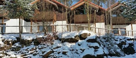 A snow-covered landscape with a wooden cabin-style building, a rocky outcrop, and a clear sky.