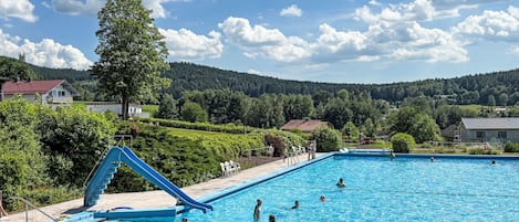En swimmingpool med rutsjebane, omgivet af grønt og huse, under en klar himmel.