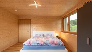 A bedroom with a wooden ceiling, a bed with a blue mattress and plaid bedding, a nightstand with a lamp, and a window with a view of greenery. - Ruhig mit einem herrlichen Blick auf die Berge. by Interhome (Schwarzenberg)
