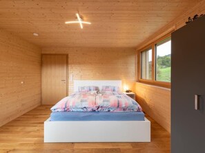 Ein Schlafzimmer mit Holzboden, einem Bett mit blauer Matratze und karierter Bettwäsche, einem Nachttisch mit Lampe und einem Fenster mit Blick auf Grünflächen.