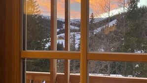Eine Holzhütte mit einem großen Fenster, das auf eine verschneite Landschaft und einen Sonnenuntergang blickt.