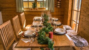 Ein Interieur einer Holzhütte zeigt einen Esstisch, gedeckt für vier Personen, mit Holzstühlen davor und ein Fenster mit Vorhängen.