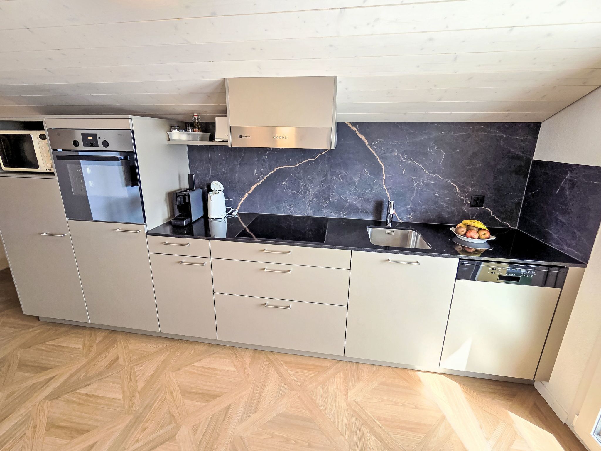 Une cuisine moderne dotée d’un four, d’un micro-ondes et d’un lave-vaisselle encastrés, d’un pare-feu en marbre noir et d’un plancher en bois disposé en motif chevrons.