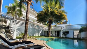 Un espace piscine avec des chaises longues, des palmiers et un bâtiment à la façade blanche.