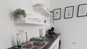 Une cuisine moderne avec des meubles blancs, un plan de travail noir et un évier en acier inoxydable. On peut voir des certificats encadrés accrochés au mur et une petite plante en pot posée sur l’étagère.