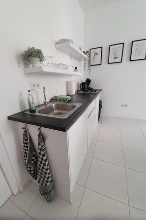 Eine moderne Küche mit weißen Schränken, einer schwarzen Arbeitsplatte und einem Edelstahlspülbecken. An der Wand befinden sich gerahmte Zertifikate und auf dem Regal ein kleiner Topfpflanze.