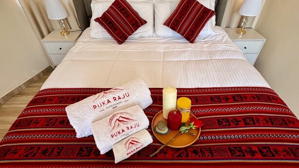 A neatly made bed with white linens and red pillows, a red and white patterned bedspread, and a tray with towels, candles, and a rose.
