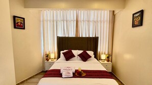 A hotel room with a large bed, white curtains, two bedside tables with lamps, and framed pictures on the walls.