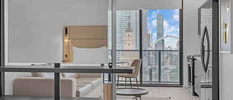 Habitación de hotel moderna con cama, sofá, una mesita y una silla. Amplia ventana con vistas a un paisaje urbano.