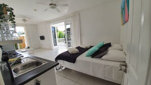 Ein kleines Zimmer mit Bett, einer Kochnische, einem Deckenventilator und einer Glasraumtür, die auf einen Balkon führt.