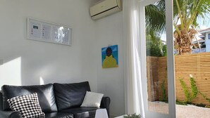 Ein modernes Wohnzimmer mit einer schwarzen Ledersofa, einem weißen Couchtisch und einer kleinen Topfpflanze. An der Wand hängt ein Gemälde, und über dem Sofa befindet sich eine Klimaanlage.
