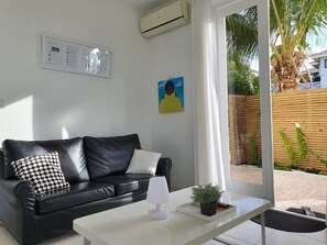 Ein modernes Wohnzimmer mit einer schwarzen Ledersofa, einem weißen Couchtisch und einer kleinen Topfpflanze. An der Wand hängt ein Gemälde, und über dem Sofa befindet sich eine Klimaanlage.