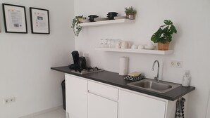 Eine moderne Küche mit weißen Schränken, einer schwarzen Arbeitsplatte, einem Spülbecken und einem Herd. An der Wand hängen zwei gerahmte Zertifikate, und auf Regalen stehen Zimmerpflanzen.