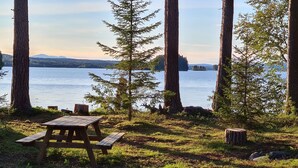 Piknikpöytä metsäisellä alueella, taustalla järvi ja vuoret.