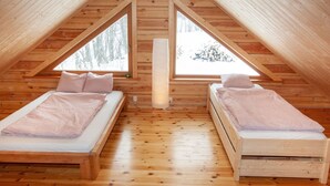Kodikas ullakkohuone, jossa on kaksi sänkyä, puulattiat ja suuret ikkunat, joista avautuu näkymä puihin.