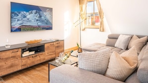 A modern living room with a large flat-screen TV mounted on the wall, a wooden cabinet with drawers, a grey sofa with multiple cushions, and a floor lamp. - Appartement Lee by Interhome (Mauterndorf)