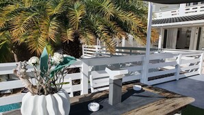 Una mesa de madera con un jarrón blanco lleno de flores, una cerca blanca y un edificio al fondo.