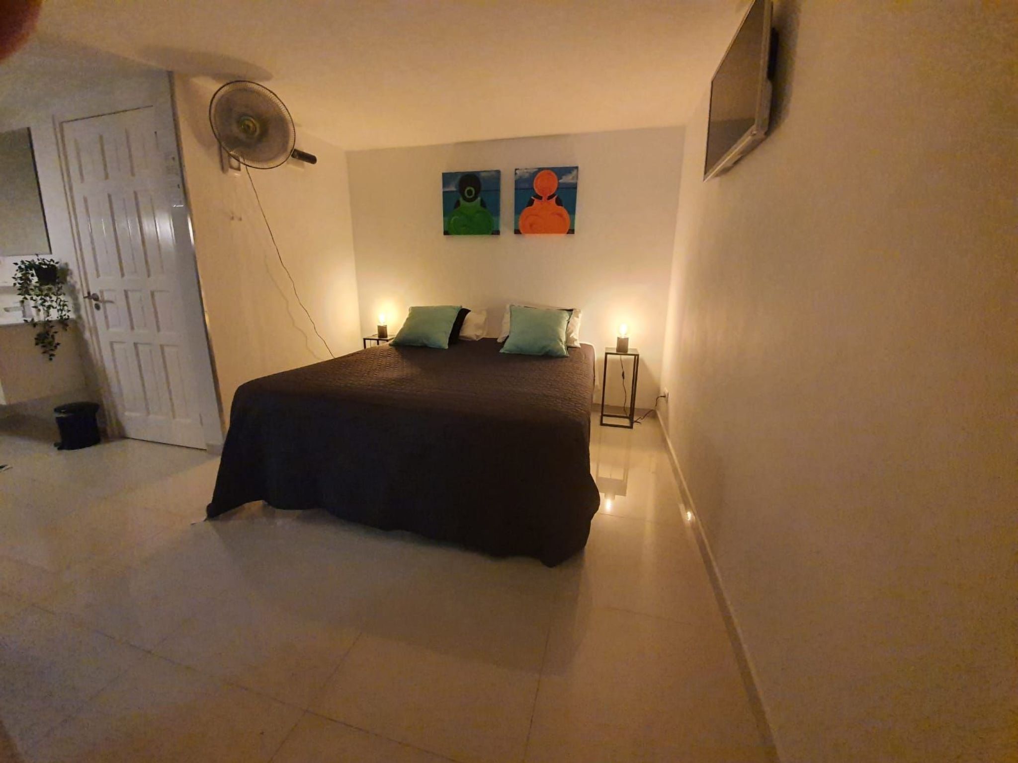 Ein Schlafzimmer mit einem Bett, zwei Nachttischen mit Lampen, einem Wandventilator und zwei Bildern.