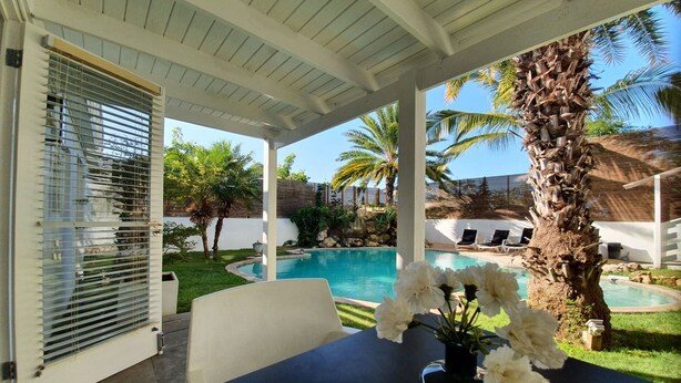 Un espacio exterior techado con piscina, palmeras y una mesa con un jarrón de flores.