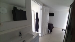 Un baño moderno con un lavamanos y un espejo blancos, una puerta que da a un dormitorio y una pequeña mesa con un mantel negro.