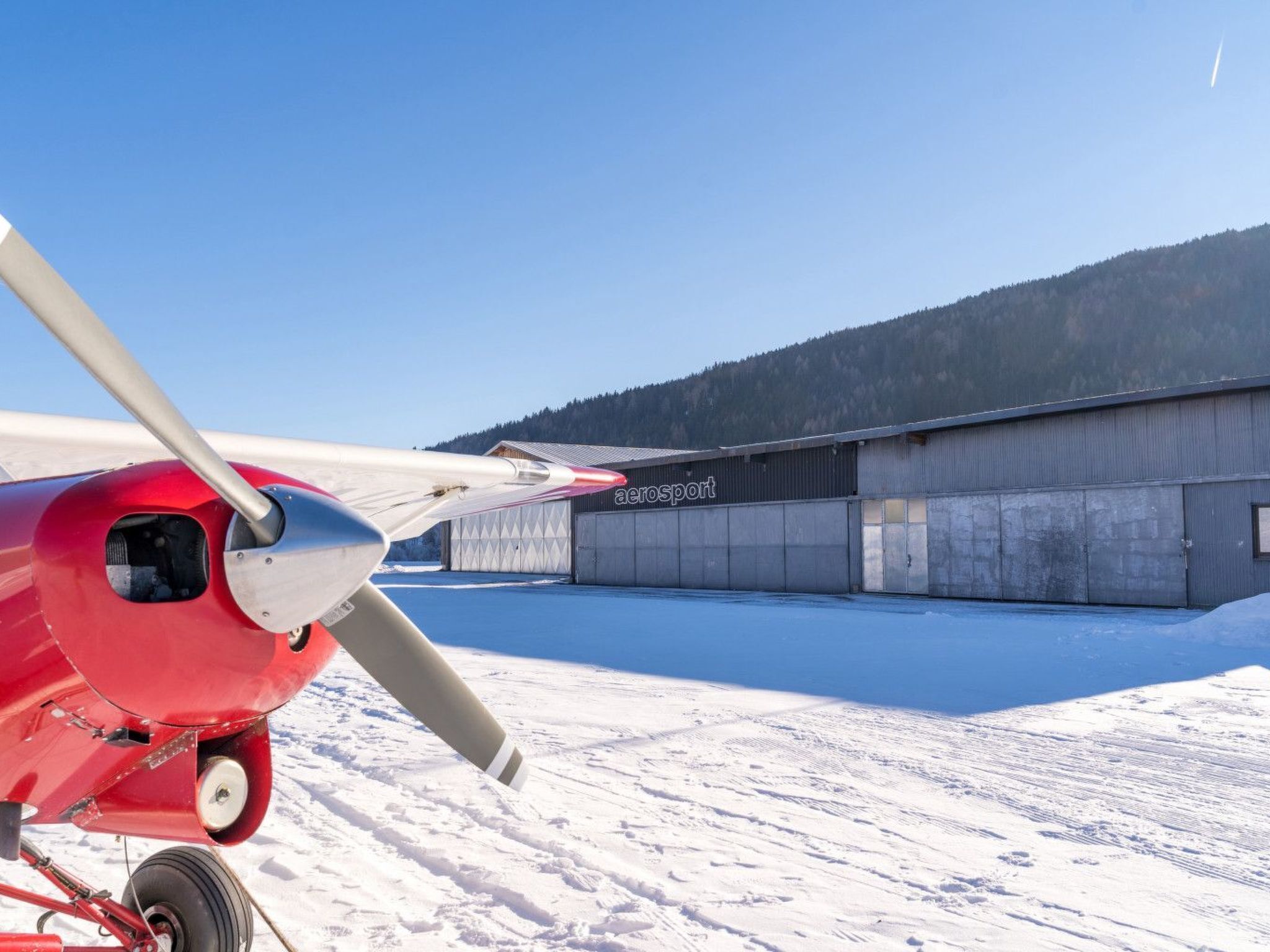 A red propeller plane parked on a snow-covered ground with a hangar labeled "aerosport" in the background.