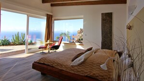 Une chambre moderne avec un grand lit, des poutres en bois, une vue sur l’océan et un coin repas comprenant une table et des chaises.