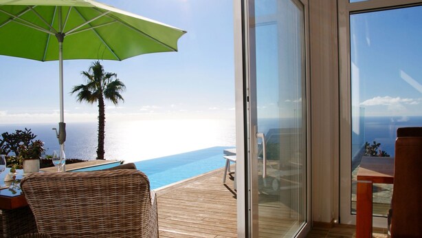Un balcon avec vue sur l’océan, une chaise en osier, une table avec un verre et un parasol vert.