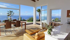 Un vaste espace de vie doté de grandes fenêtres offrant une vue sur l’océan, d’une table à manger en bois et d’un coin salon confortable.