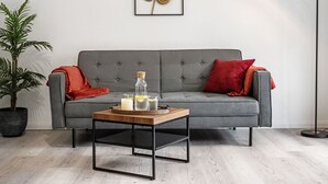 Een moderne woonkamer met een grijze bank, een klein houten bijzettafeltje, een vloerlamp en een plant in pot.