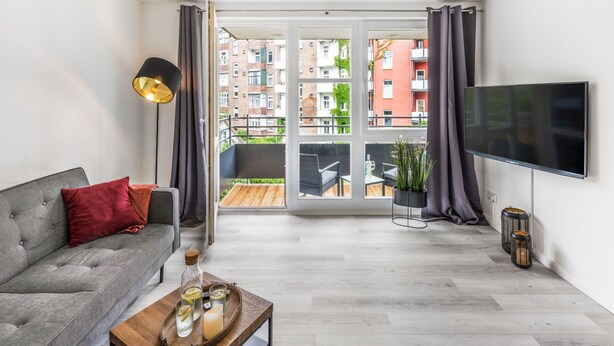 Een moderne woonkamer met een grijze bank, een salontafel met drankjes, een televisie en een balkon met uitzicht op gebouwen.