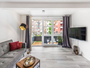 Un soggiorno moderno con un divano grigio, un tavolino con bevande, una TV e un balcone con vista sugli edifici.