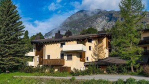 Ein mehrstöckiges Gebäude mit geneigtem Dach, umgeben von Bäumen und Bergen im Hintergrund.